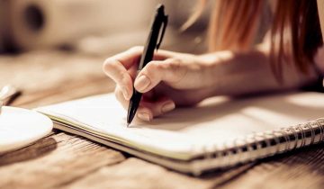 Close up view of womans hand writing with pen on notebook on wooden table.